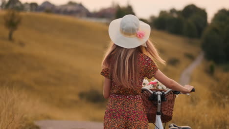 Von-Hinten-Dreht-Sich-Ein-Fröhliches-Blondes-Mädchen-In-Kleid-Und-Hut-Um,-Lächelt-Fröhlich,-Schaut-In-Die-Kamera-Und-Flirtet,-Während-Sie-Im-Sommer-Mit-Fahrrad-Und-Blumen-über-Das-Feld-Schlendert.